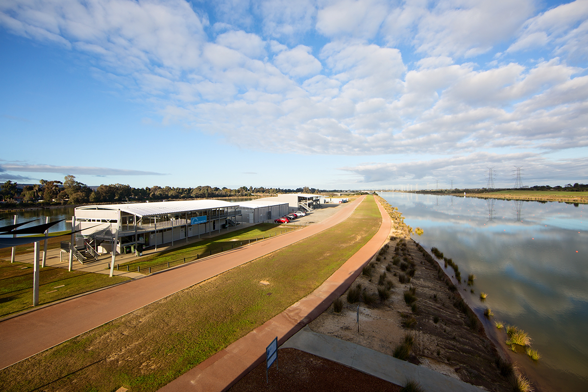 A elevated angle of a cycle path running parallel to the water and main building at the Champion Lakes Regatta Centre