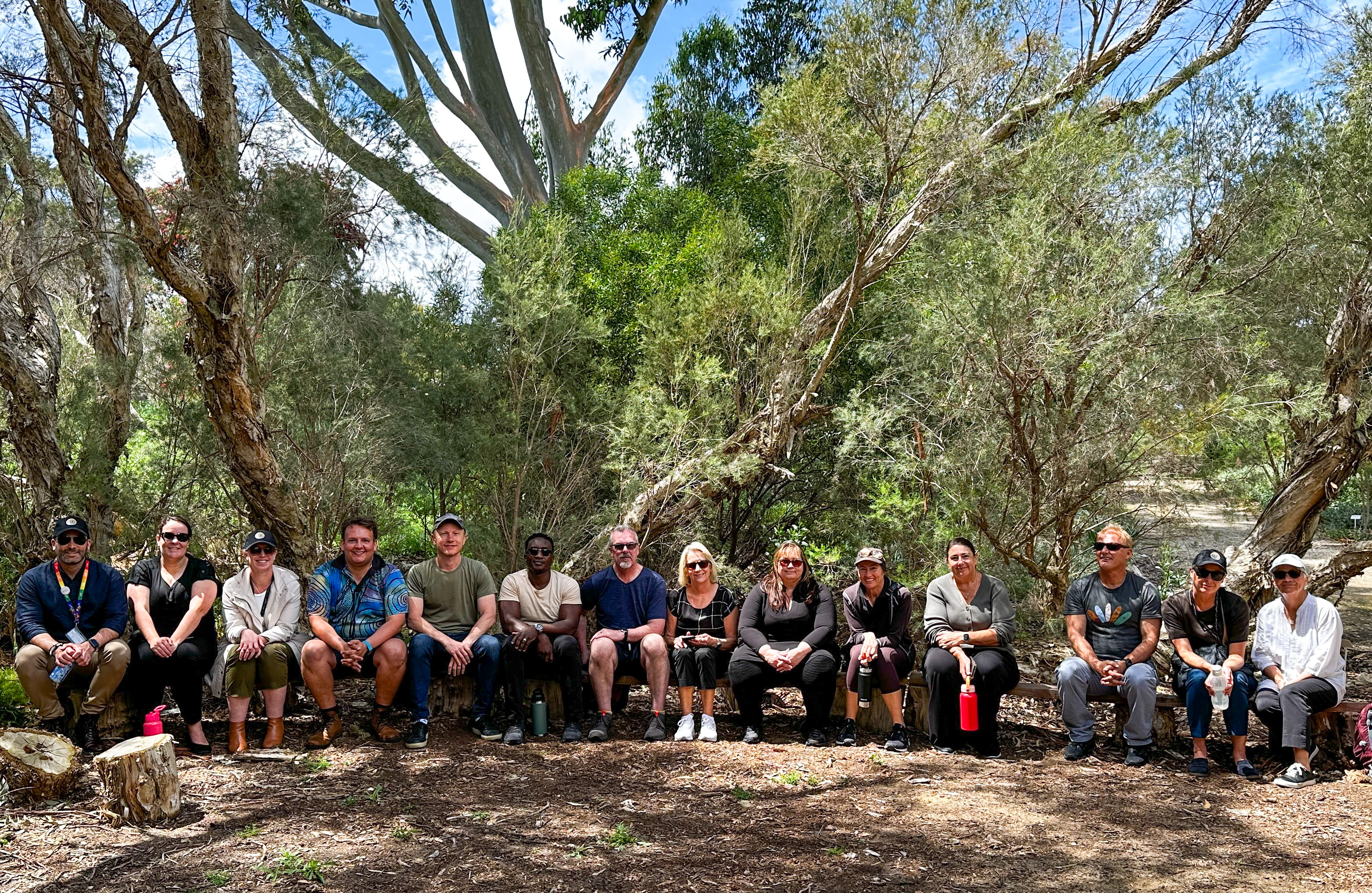 A group photo of multiple people sitting together in front of trees and greenery