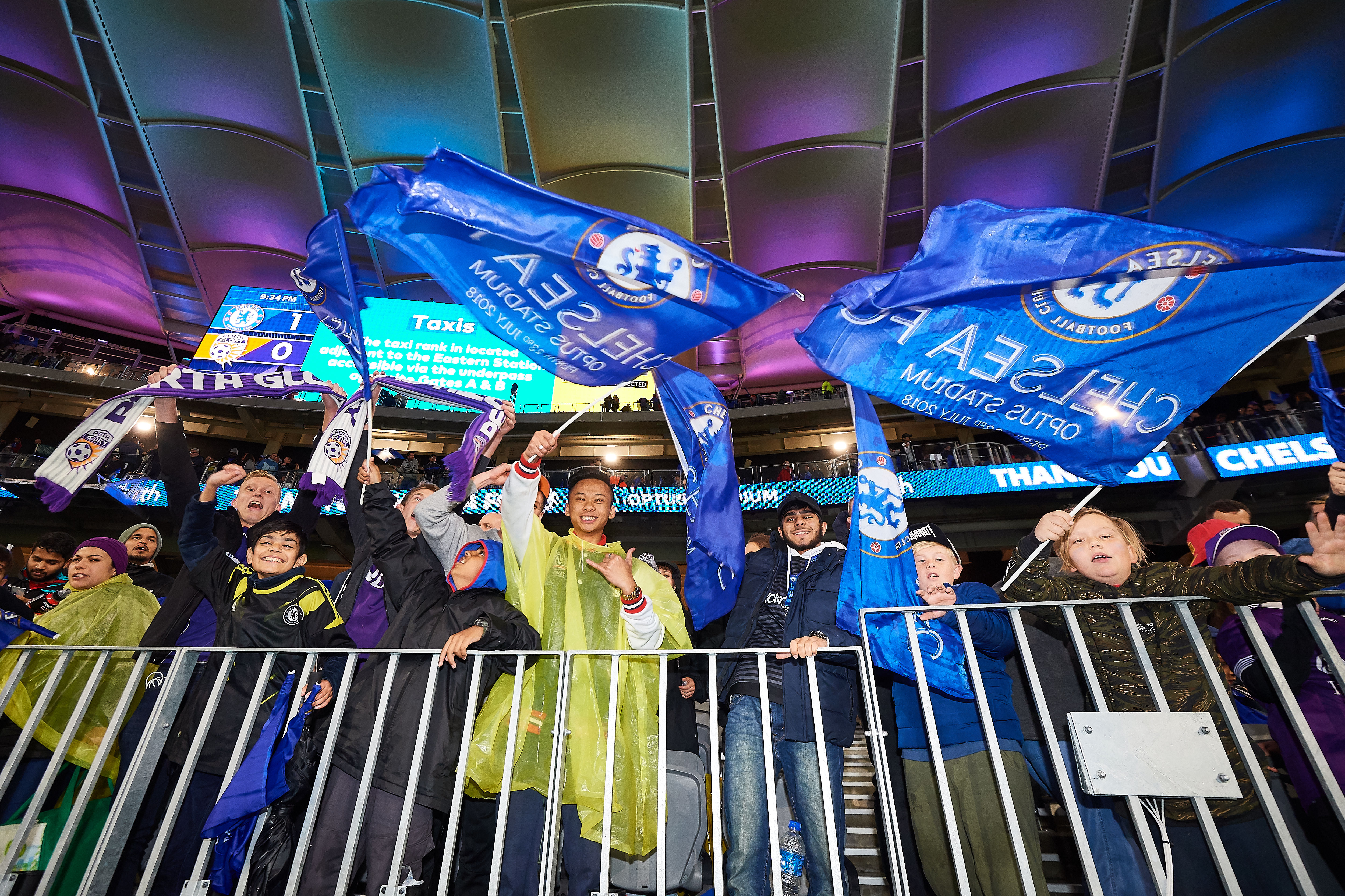 A photo of Chelsea Football Club fans waving flags and celebrating during a game at Optus Stadium
