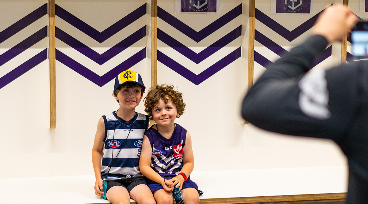 A photo of two children wearing AFL guernseys having their picture taken in the change rooms at Optus Stadium