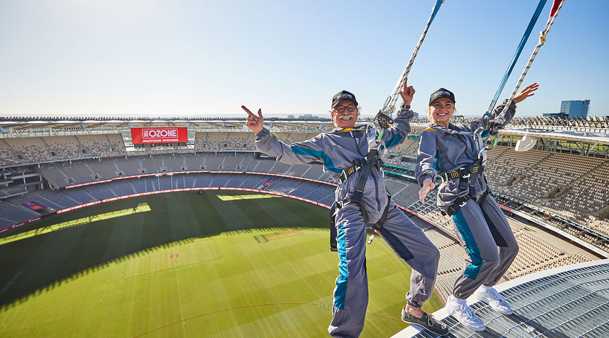 A photo of two people, secured by harness, standing on the edge of the Optus Stadium roof