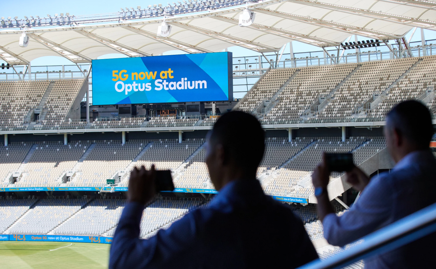 Photo from behind two people with focus on the Optus Stadium big screen in the background
