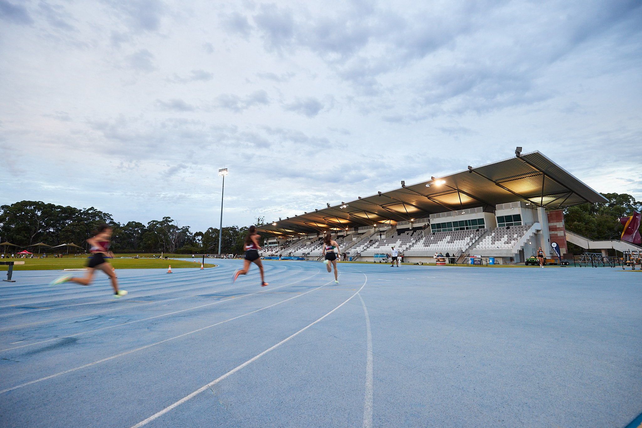 A photo of three runners rounding a corner of track near the grandstand seating area at WA Athletics Stadium
